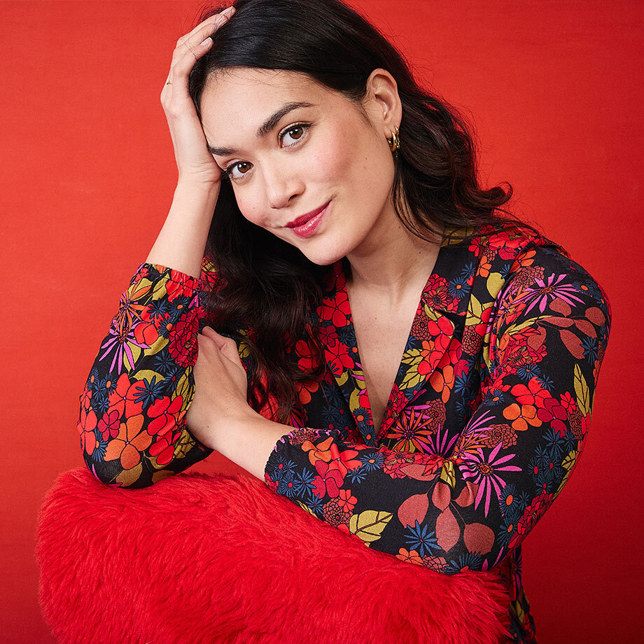 Femme brune souriante portant une robe &agrave; imprim&eacute; floral color&eacute;, appuy&eacute;e sur un coussin rouge duveteux devant un fond rouge vif.