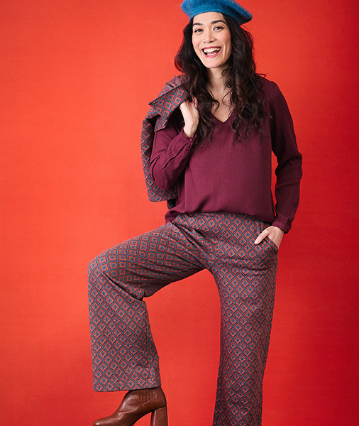 femme-souriante-portant-un-beret-bleu-une-blouse-bordeaux-et-un-pantalon-a-motif-geometrique-bleu-et-rouge-posant-sur-un-fond-rouge-orange-