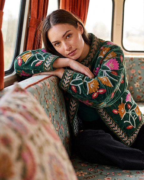 femme-assise-dans-une-banquette-de-train-au-decor-retro-appuyee-sur-ses-bras-portant-une-veste-verte-brodee-de-motifs-floraux-colores-