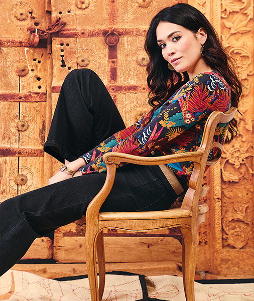 une-femme-aux-longs-cheveux-bruns-ondules-est-assise-sur-une-chaise-en-bois-sculpte-posant-devant-une-porte-en-bois-ancienne-et-ornee--elle-porte-un-haut-a-manches-longues-avec-un-motif-floral-tropical-et-un-pantalon-noir-large--elle-regarde-directement-l-appareil-photo-avec-un-leger-sourire-son-bras-pose-sur-l-accoudoir-de-la-chaise-