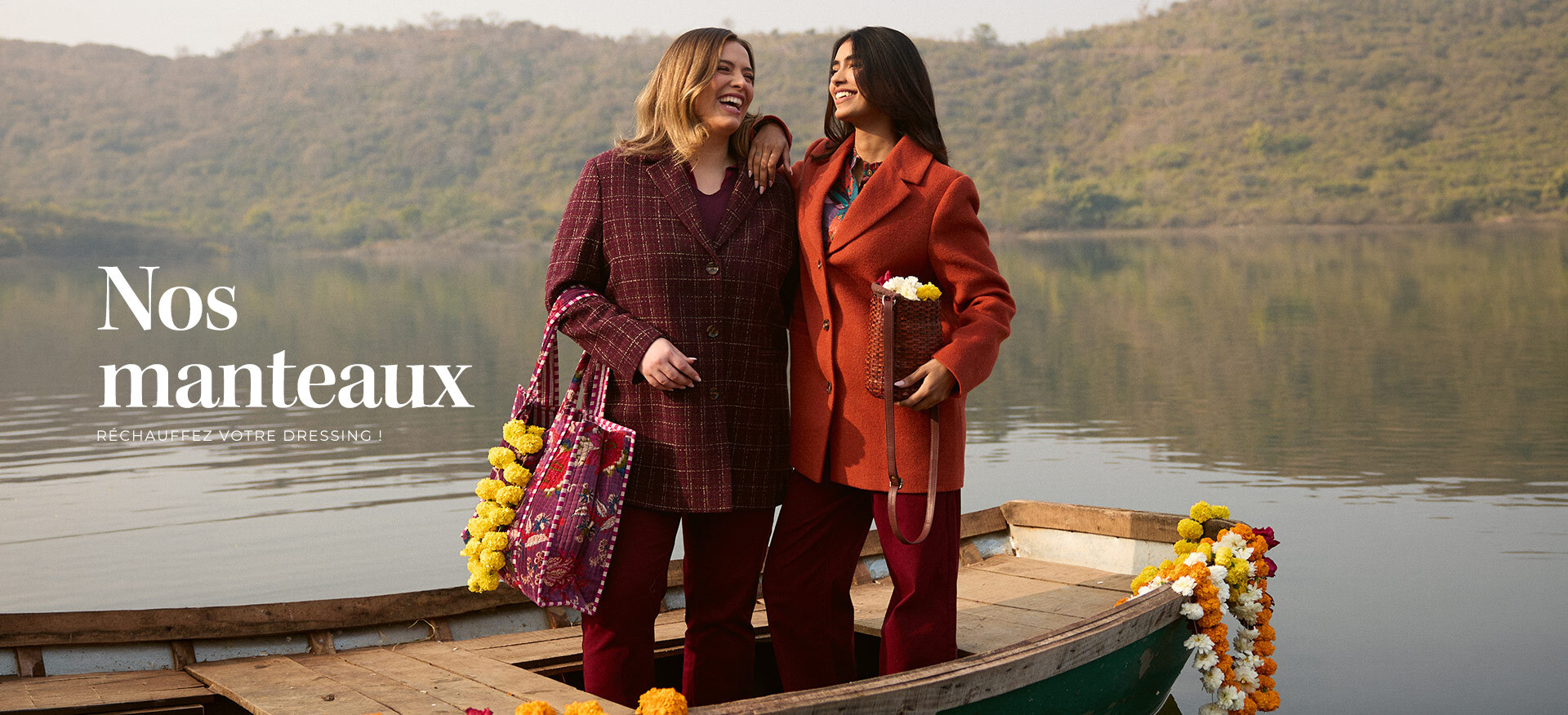 deux-femmes-souriantes-portant-des-manteaux-d-hiver--un-manteau-a-carreaux-bordeaux-et-un-manteau-orange--se-tiennent-sur-une-barque-en-bois-entouree-d-eau-et-de-collines--texte-superpose---nos-manteaux---rechauffez-votre-dressing--
