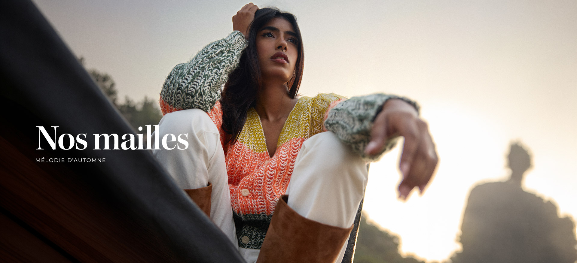 mannequin-assise-en-exterieur-au-coucher-du-soleil-portant-un-cardigan-en-grosse-maille-multicolore-jaune-corail-et-vert-associe-a-un-pantalon-blanc-et-des-bottes-camel--collection--nos-mailles--melodie-dautomne