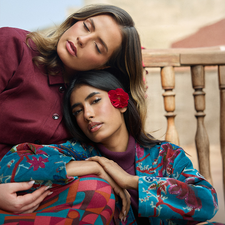 Deux femmes souriantes photographi&eacute;es en contre-plong&eacute;e, v&ecirc;tues de tenues color&eacute;es aux imprim&eacute;s graphiques et floraux, l&rsquo;une tenant des fleurs rouges au premier plan.